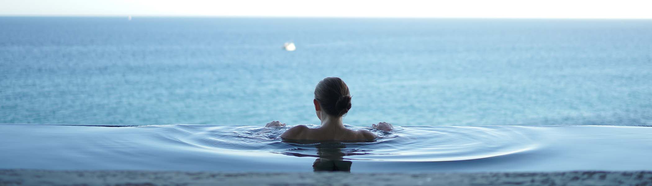 The back of a woman in an infinity pool looking out at the sea.
